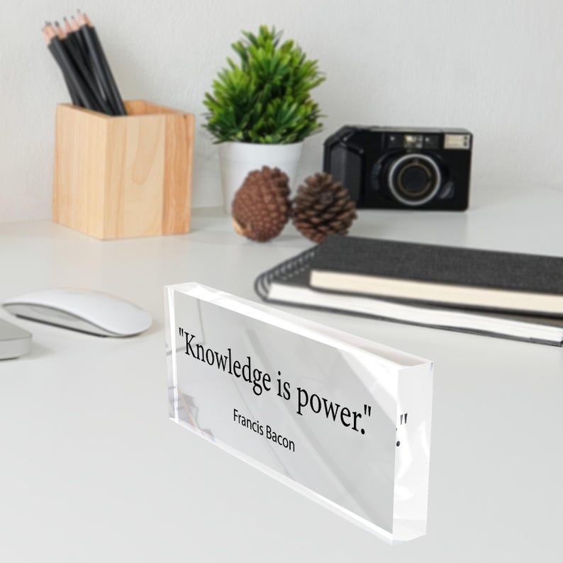 May include: A clear acrylic block with the quote "Knowledge is power." by Francis Bacon. The block is on a white surface, with a wooden pencil holder, a small plant, and a camera in the background.
