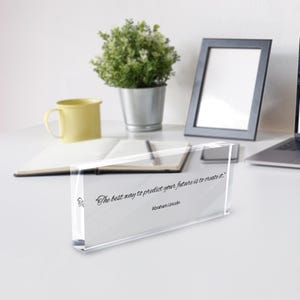 May include: A clear acrylic block with the quote "The best way to predict your future is to create it." by Abraham Lincoln. The block is on a white surface with a yellow mug, a small potted plant, and a picture frame in the background.