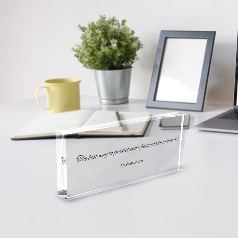 May include: A clear acrylic block with the quote "The best way to predict your future is to create it." by Abraham Lincoln. The block is on a white surface with a yellow mug, a small potted plant, and a picture frame in the background.