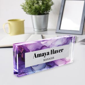 May include: A clear acrylic desk nameplate with a purple and blue abstract design. The name "Amaya Haver" and the title "MANAGER" are printed in black text.