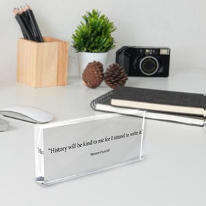 May include: A clear acrylic block displays a quote by Winston Churchill: "History will be kind to me for I intend to write it." The block sits on a white desk with a wooden pencil holder, a camera, and a small potted plant.