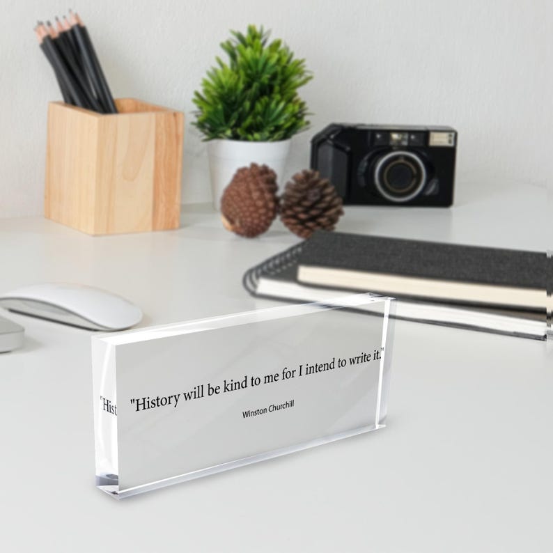 May include: A clear acrylic block displays a quote by Winston Churchill: "History will be kind to me for I intend to write it." The block sits on a white desk with a wooden pencil holder, a camera, and a small potted plant.