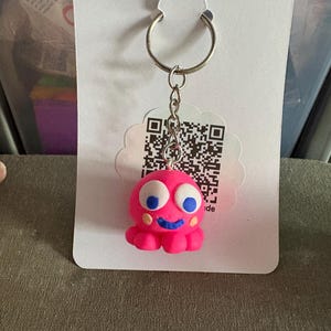 May include: Pink octopus keyring with blue eyes and a smile. The keyring has a silver ring and a QR code on the back.