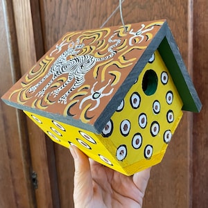 May include: A yellow and brown birdhouse with a black roof and a painted tiger design. The birdhouse has a round hole for birds to enter.