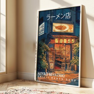 May include: A framed art print depicting a Japanese ramen shop at night. The artwork features a detailed illustration of the shop's exterior, with lanterns, signage in Japanese, and the text "HAYAO MIYAZAKI GHIBLI RAMEN SHOP".