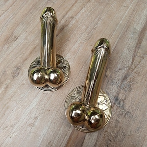 Puede incluir: Dos manijas de puerta de color latón, cada una con la forma de un falo estilizado. Las manijas están montadas sobre placas traseras circulares y ornamentadas con un diseño floral. El fondo es una superficie de madera clara.