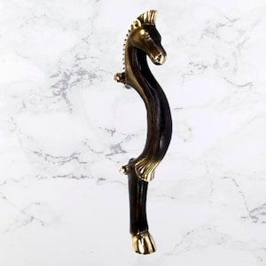 May include: A bronze-toned door handle shaped like a horse. The handle features a horse head at the top and a curved body. The horse's mane and details are highlighted with a gold finish. The handle is designed to be mounted on a door.