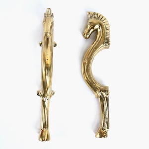 Könnte beinhalten: Zwei messingfarbene Schrankgriffe, jeweils in Form eines Pferdes. Einer ist ein gerader Griff mit einem Pferdekopf oben. Der andere ist ein gebogener Griff mit einem Pferdekopf. Beide haben detaillierte Merkmale.