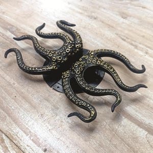 May include: A black metal octopus wall decor with gold accents. The octopus has eight tentacles with a textured surface.