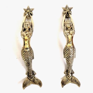 May include: Two bronze-coloured mermaid figurines, each holding a star above their heads. The mermaids have long hair and detailed fishtails. These decorative figurines are likely for a nautical or fantasy-themed setting.