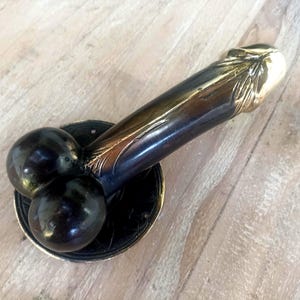 Puede incluir: Un tirador de puerta de tono bronce con un diseño único. El tirador presenta una forma cilíndrica con detalles decorativos y una base circular con tres elementos esféricos. El tirador está montado sobre una superficie de madera.