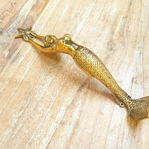 May include: A gold-colored mermaid figurine holding a starfish. The mermaid has detailed scales and flowing hair. The figurine is made of metal and is likely a decorative item.