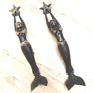 Peut inclure: Deux figurines de sirènes en bronze, chacune tenant une étoile de mer au-dessus de sa tête. Les sirènes ont des corps noirs avec des accents dorés sur leurs hauts en coquillages et leurs ceintures. Leurs queues sont détaillées avec des écailles, et les étoiles de mer sont également en bronze.