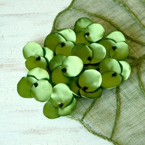 May include: A cluster of green satin flower petals with black centers, arranged on a green woven leaf-shaped background.