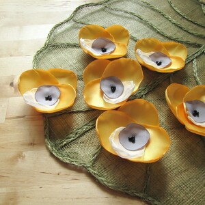 May include: Seven yellow fabric flowers with black centers arranged on a green and brown textured surface.