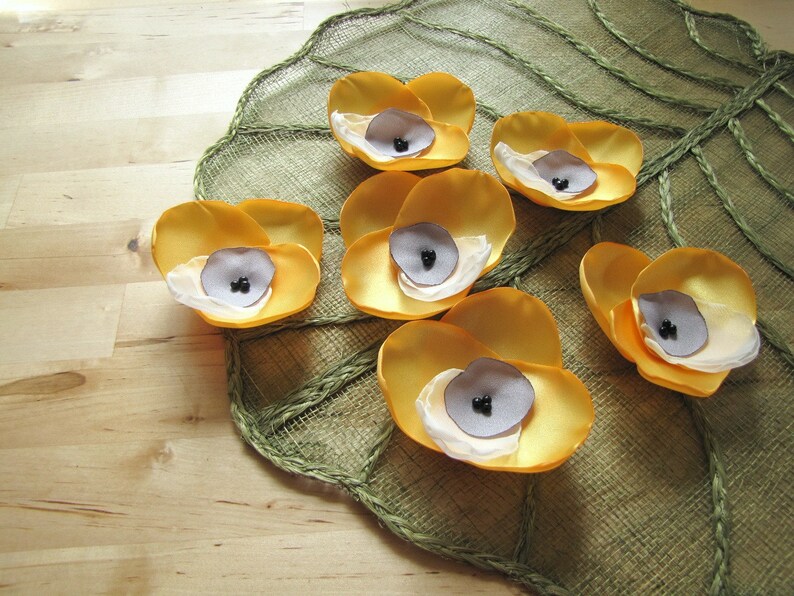 May include: Seven yellow fabric flowers with black centers arranged on a green and brown textured surface.