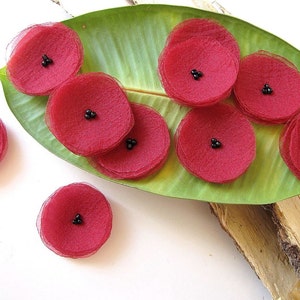 May include: A collection of ten red fabric flower embellishments with black bead centres. The flowers are arranged on a green leaf and a piece of wood.