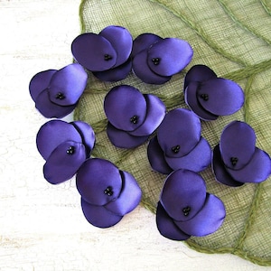 May include: Purple fabric flower embellishments with black centers. The flowers are arranged on a green textured surface.