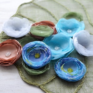 May include: Assortment of fabric flowers in various colors and patterns, including blue, green, white, and brown. The flowers are made of layered fabric and have a delicate, handmade appearance.