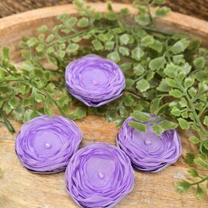 May include: Four lavender fabric flower embellishments with a beaded center, arranged on a wooden tray with green foliage. The flowers have a layered petal design and are a soft, pastel purple color.