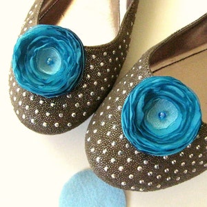 Puede incluir: Un par de pisos marrones con un acabado brillante y una flor de tela azul turquesa como adorno en cada zapato.
