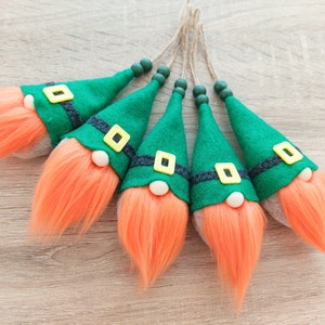 May include: Five green felt gnome ornaments with orange yarn hair and black and yellow felt accents. The gnomes are hanging from a string.