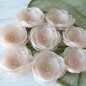 May include: Eight blush pink silk flower embellishments with a pearl center. The flowers are arranged on a green textured surface.