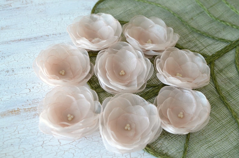 May include: Eight blush pink silk flower embellishments with a pearl center. The flowers are arranged on a green textured surface.