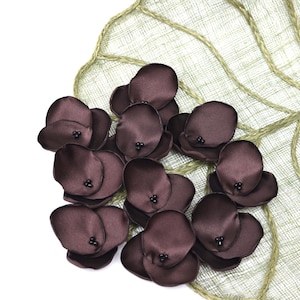 May include: Dark brown satin flower petals with black bead centres, arranged on a light green, leaf-shaped fabric with a woven texture. The petals create a decorative floral arrangement.