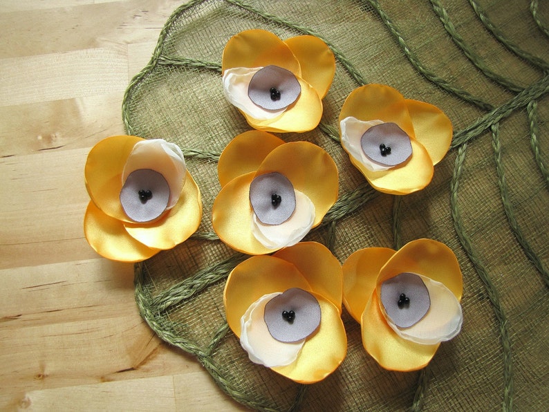 May include: Six yellow fabric flowers with white centers and black beads in the center of each flower. The flowers are arranged on a green and brown woven fabric background.