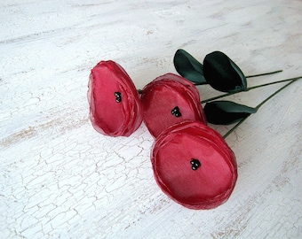 May include: Three red fabric flowers with black centers on a white textured background. The flowers are attached to green stems.