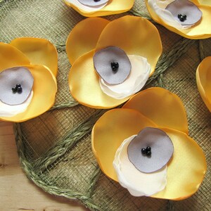 May include: A collection of six yellow fabric flower embellishments with gray centers and black beads. The flowers are arranged on a woven brown background.