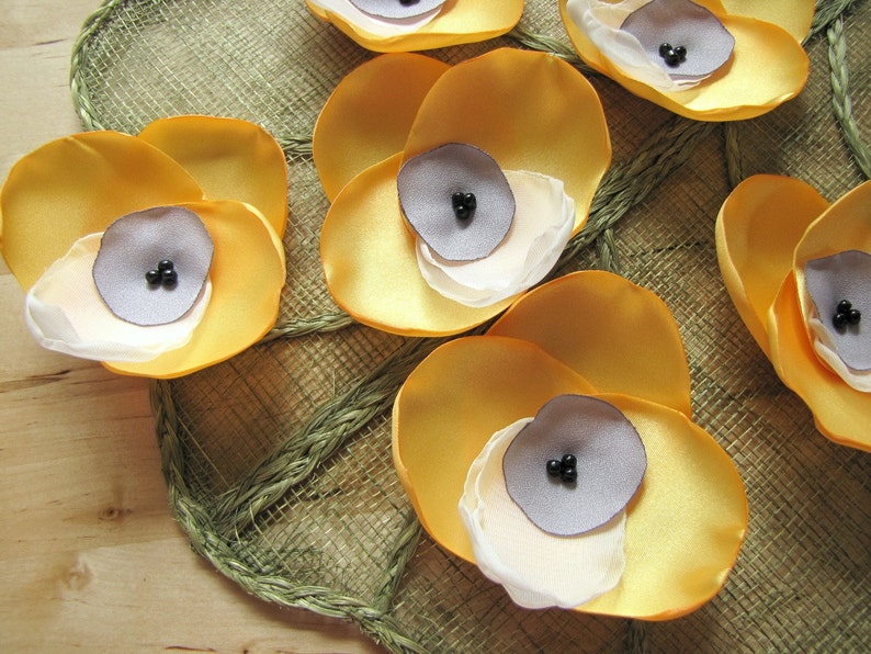 May include: A collection of six yellow fabric flower embellishments with gray centers and black beads. The flowers are arranged on a woven brown background.