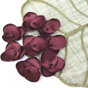 May include: A collection of burgundy satin flower petals, each with a cluster of black beads at the centre, are arranged on a light green, leaf-shaped fabric. The petals are clustered, creating a delicate floral design. The background is white.