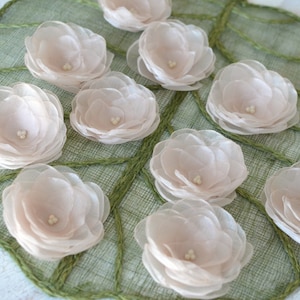 May include: A collection of ten delicate, pale pink fabric flowers arranged on a green woven surface. The flowers are made of multiple layers of sheer fabric, giving them a soft and airy appearance.