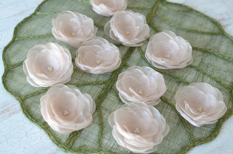 May include: A collection of ten delicate, pale pink fabric flowers arranged on a green woven surface. The flowers are made of multiple layers of sheer fabric, giving them a soft and airy appearance.