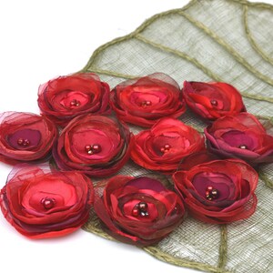 May include: A collection of red fabric flowers with dark red centers and small bead accents. The flowers are arranged on a green, leaf-shaped decorative piece with a woven texture. The background is white.