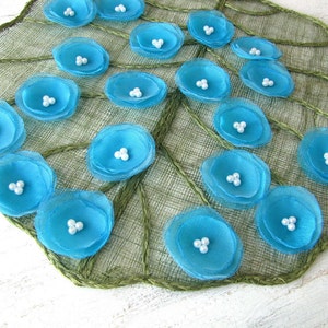May include: A close-up of a blue and white fabric flower embellishment. The flower is made of multiple layers of blue fabric with white pearl beads in the centre. The flower is on a green burlap background.