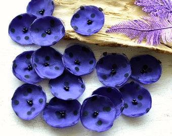 May include: A collection of purple fabric flower embellishments with black polka dots and beaded centers. The flowers are arranged on a light-colored wooden surface, with a purple glitter feather in the background. These are ideal for crafting and DIY projects.