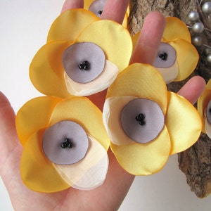 May include: Five yellow fabric flowers with a layered design. Each flower has a center made of a light purple fabric with three black beads.
