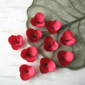 May include: A collection of ten red felt poppies with black centres. The poppies are arranged on a white wooden surface with a green leaf-shaped mat.