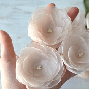 May include: Three delicate, blush-colored fabric flowers with pearl centers. The flowers are layered and have a soft, ethereal look.