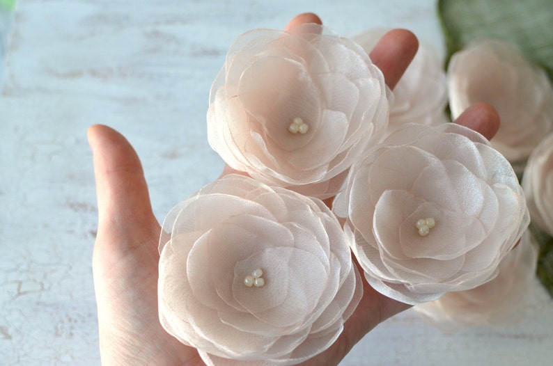 May include: Three delicate, blush-colored fabric flowers with pearl centers. The flowers are layered and have a soft, ethereal look.