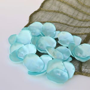 May include: A cluster of light blue satin flower petals with a subtle sheen, resting on a textured, olive green leaf-shaped fabric. The petals have a delicate, translucent quality.
