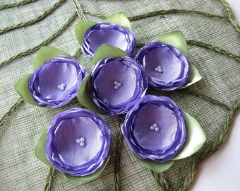 May include: Six lavender fabric flowers with green leaves and pearl accents. The flowers are arranged on a woven green background.