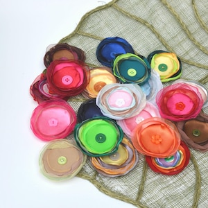 May include: A collection of colorful fabric flower embellishments in shades of pink, orange, green, and blue, each with a button center. The flowers are arranged on a textured, woven, light green background.