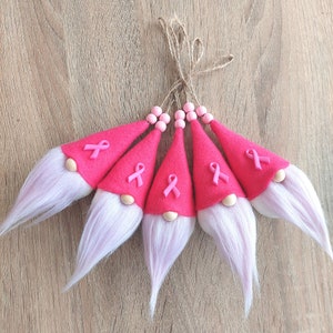 May include: Five pink felt gnomes with pink ribbon accents and wooden noses. The gnomes are tied together with twine and have long white beards.