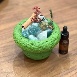 May include: A green, textured bowl filled with blue and white crystals, a cinnamon stick, and dried botanicals. An amber glass bottle with a dropper is to the right. The arrangement is on a wooden surface.