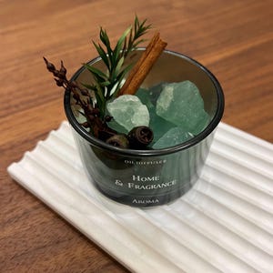 May include: A glass oil diffuser with the text "Home & Fragrance" and "Aroma". The diffuser contains green stones, a cinnamon stick, and sprigs of greenery. It sits on a white, ridged tray.