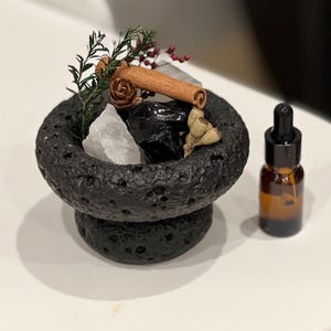 May include: A black, textured lava stone bowl filled with crystals, dried botanicals, and a cinnamon stick. A small amber glass bottle with a black dropper sits beside the bowl. The bowl is on a white surface.